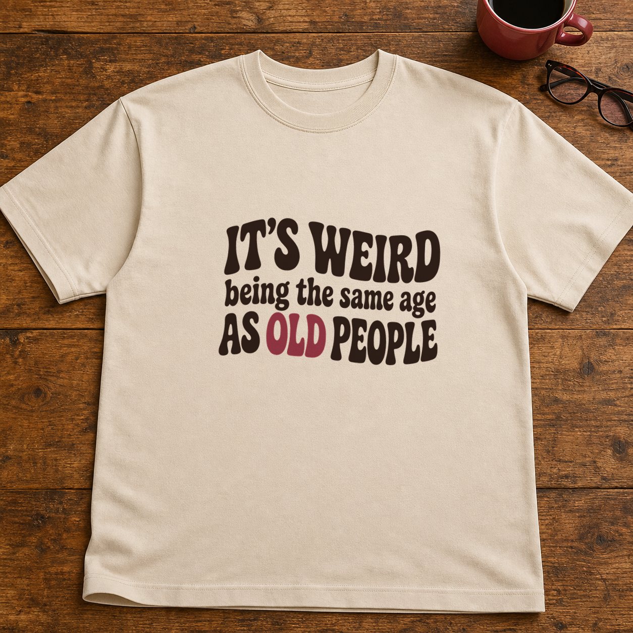 Sand tee printed with 'It's weird being the same age as old people', laying flat on a wooden table beside a raspberry coffee mug and cat-eye glasses